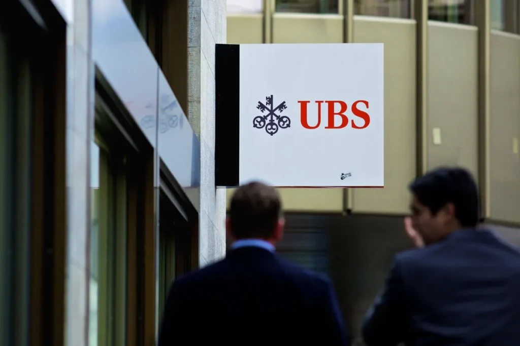 Bankers from UBS walk outside the company's offices, reflecting the ongoing job cuts and integration efforts mentioned in the article. PHOTO: Gianluca Colla/Bloomberg/100318